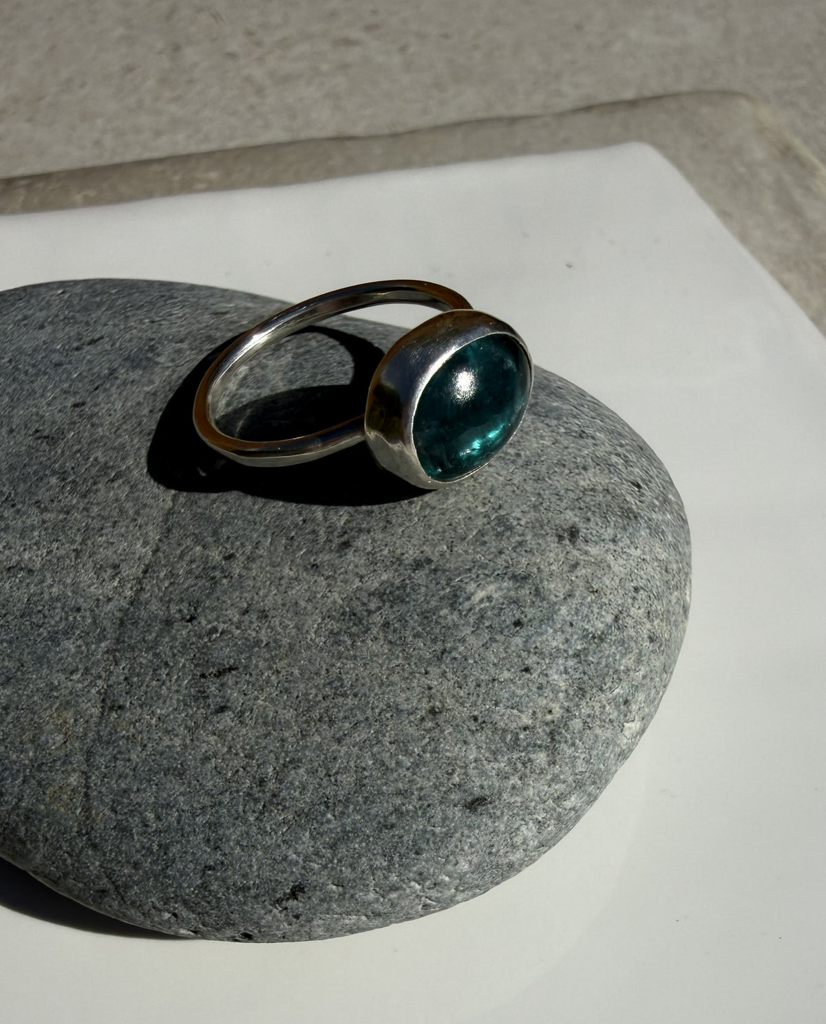 A silver ring with a round, bezel-set dark teal gemstone, displayed on a smooth gray stone against a white surface.