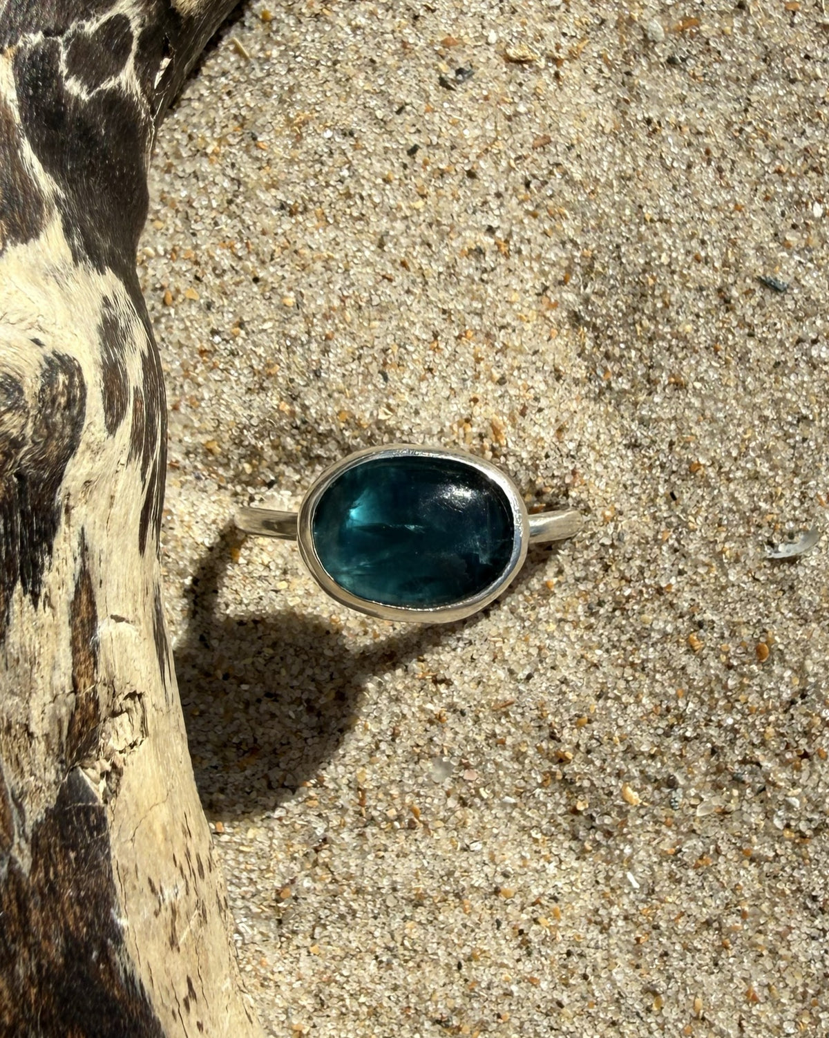 A silver ring with an oval, deep blue-green stone set in natural sand next to a piece of driftwood.