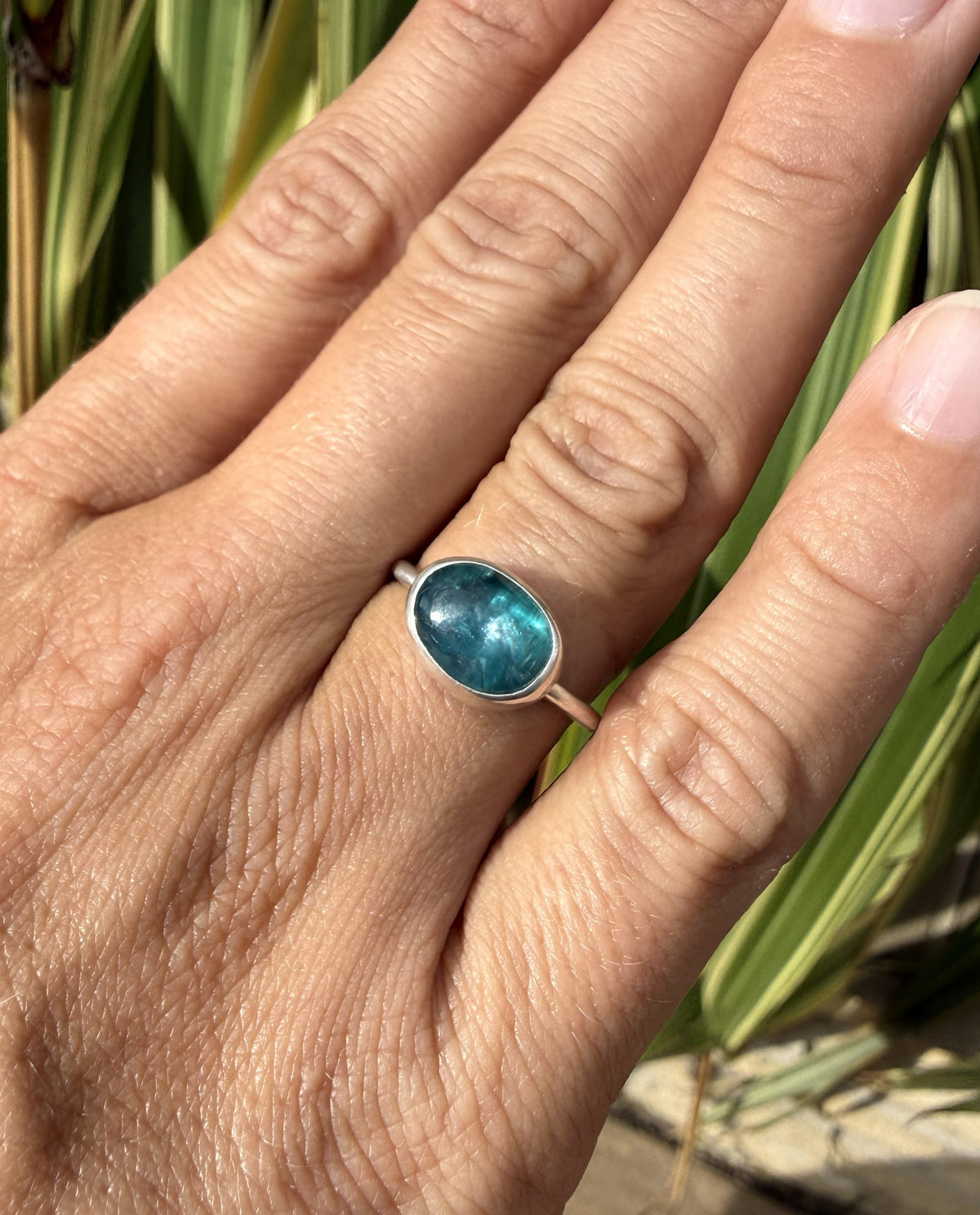 A close-up of a hand wearing a silver ring with an oval blue-green stone, set against a blurred background of green leaves.