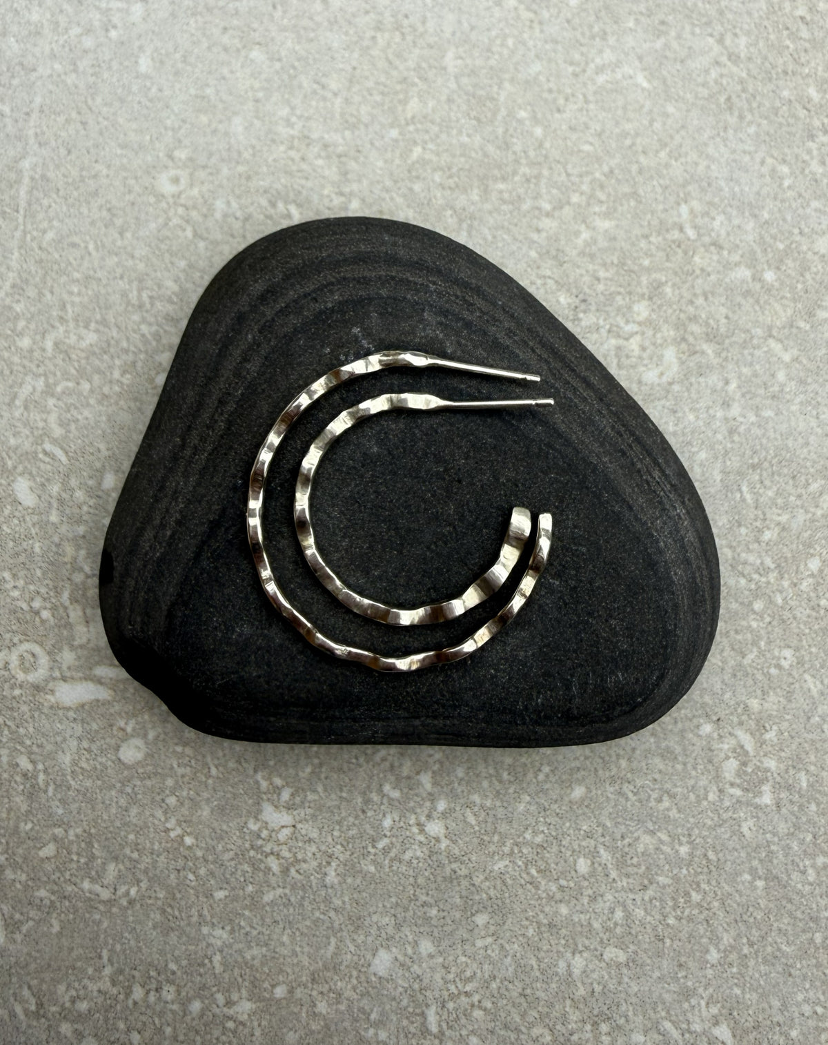 A pair of hammered silver hoop earrings displayed on a dark, textured stone against a light gray stone surface.