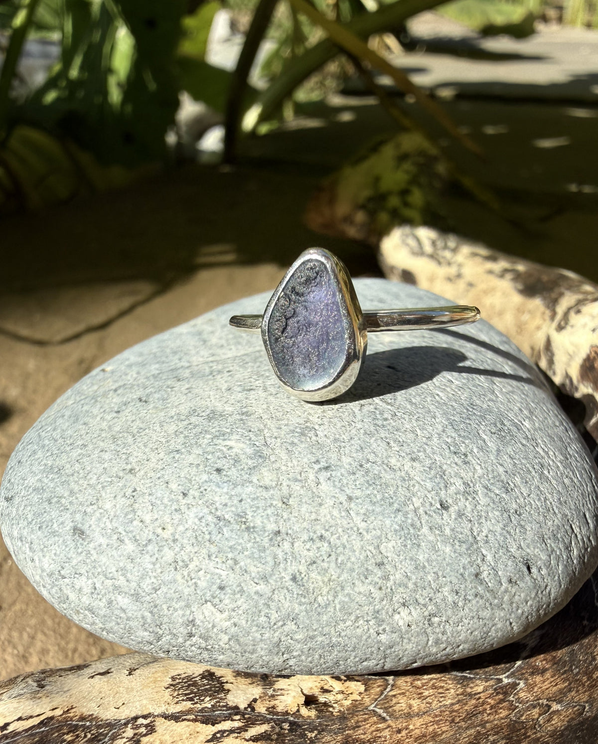 A silver ring with a teardrop-shaped lavender-purple stone resting on a smooth gray stone outdoors.