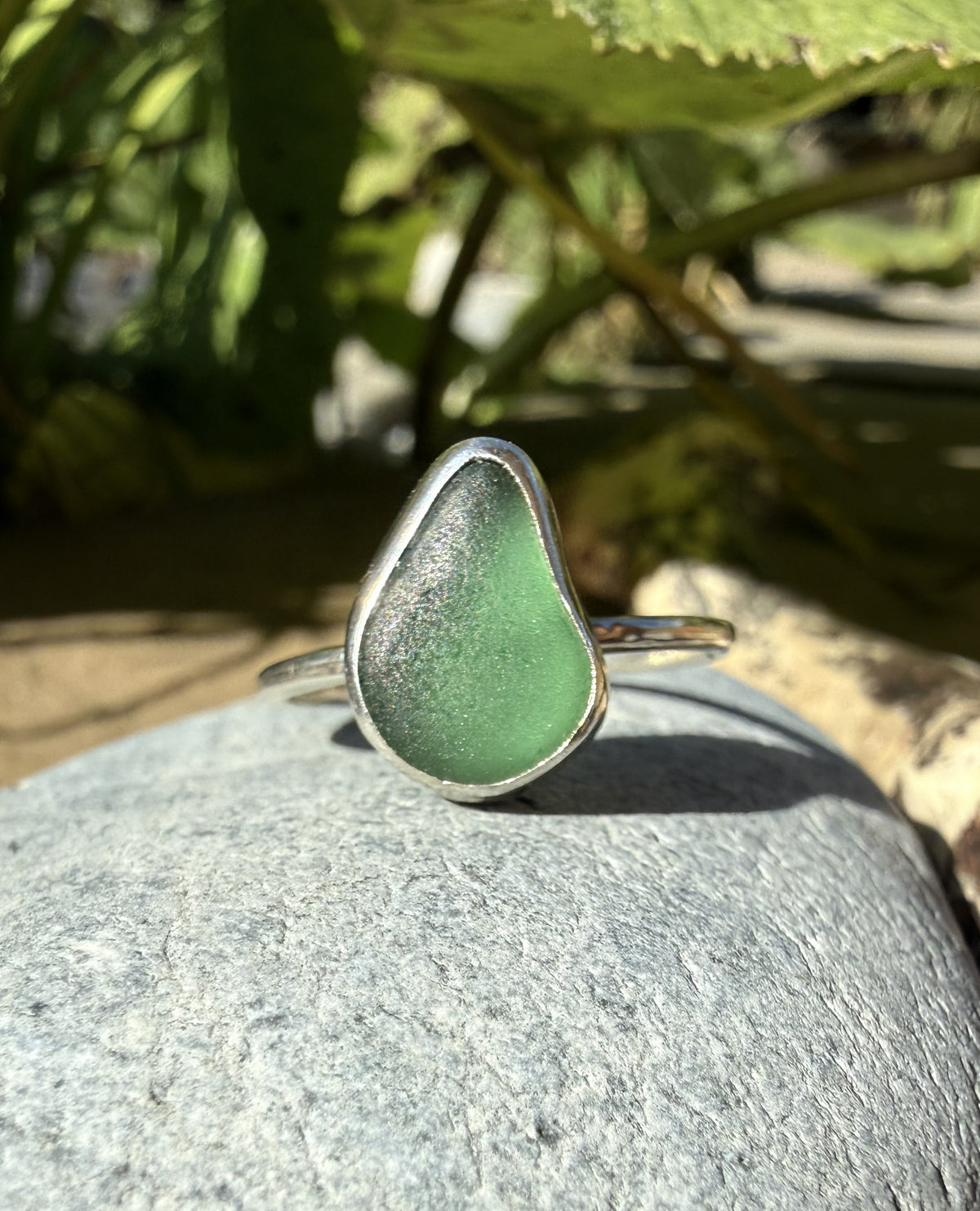 A sea glass ring with a teardrop-shaped light green stone set in a silver band, resting on a gray stone outdoors with green foliage in the background.