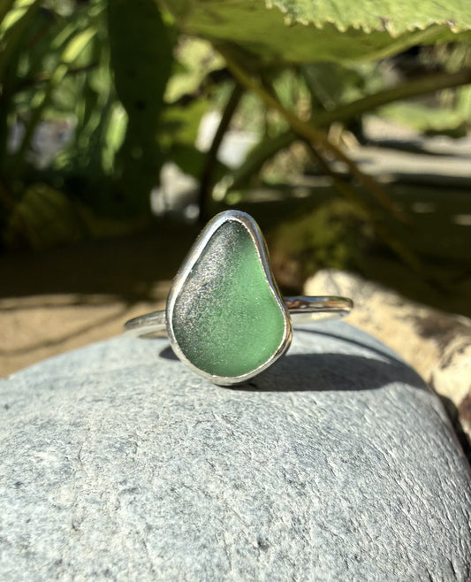 A sea glass ring with a teardrop-shaped light green stone set in a silver band, resting on a gray stone outdoors with green foliage in the background.