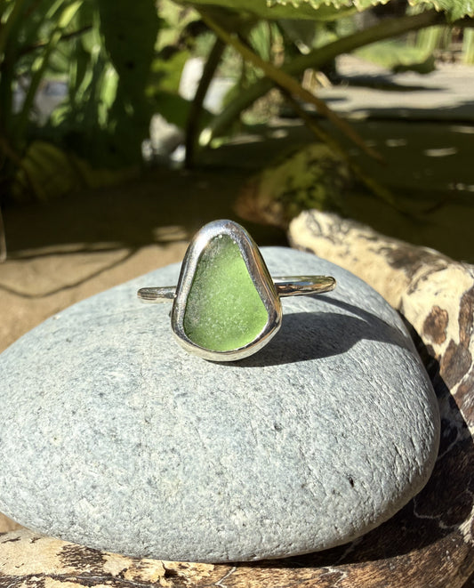 A sea glass ring with a light green teardrop-shaped stone in a silver bezel setting, displayed on a smooth gray stone outdoors with blurred green foliage in the background.