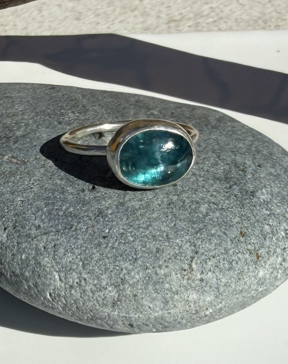 A silver ring with an oval-cut teal gemstone resting on a smooth gray stone surface.