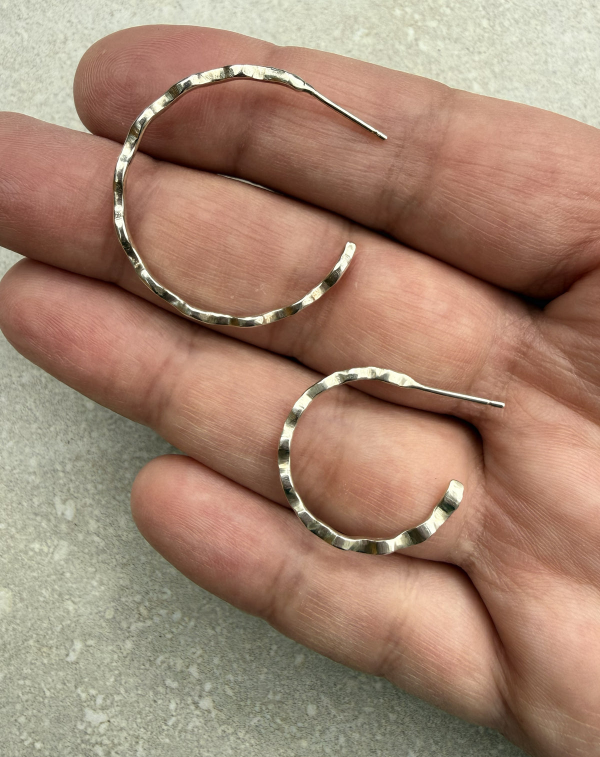 A pair of hammered silver hoop earrings held in a person's hand against a light gray stone surface.