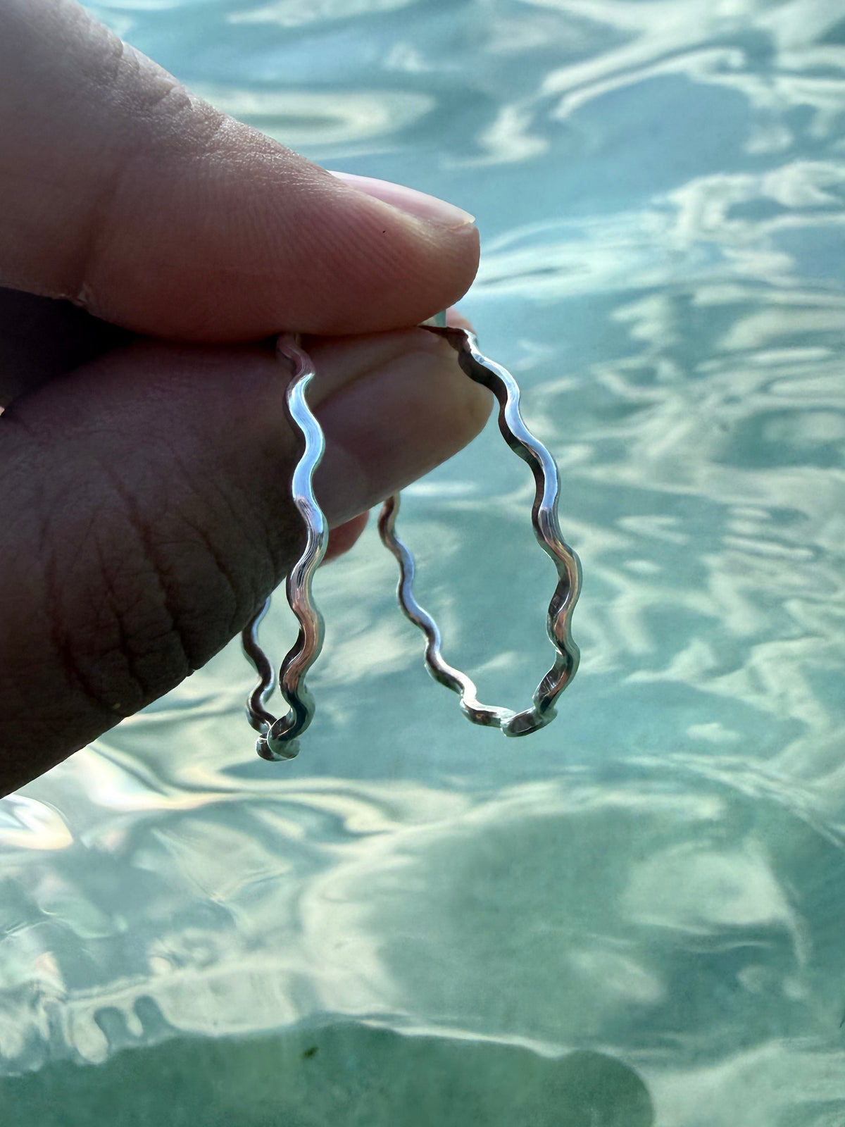 A close-up of a hand holding two thin, wavy silver hoop earrings against a background of clear, sunlit water.